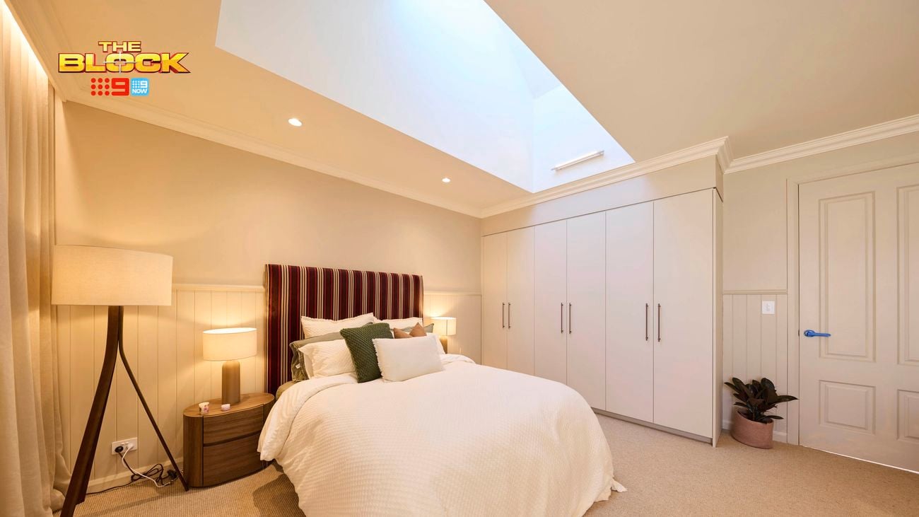 Cream bedroom with a double bed and a patterned bedhead, white linens and a lamp in the corner and a wardrobe opposite.
