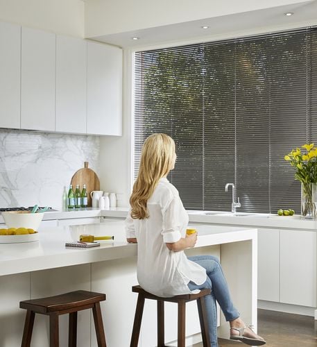 Young woman in a modern kitchen looking out a window with grey venetian blinds