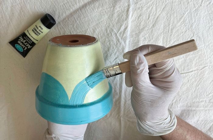 Hands wearing gloves paint a terracotta pot with light yellow and blue acrylic paint on a fabric-covered surface.