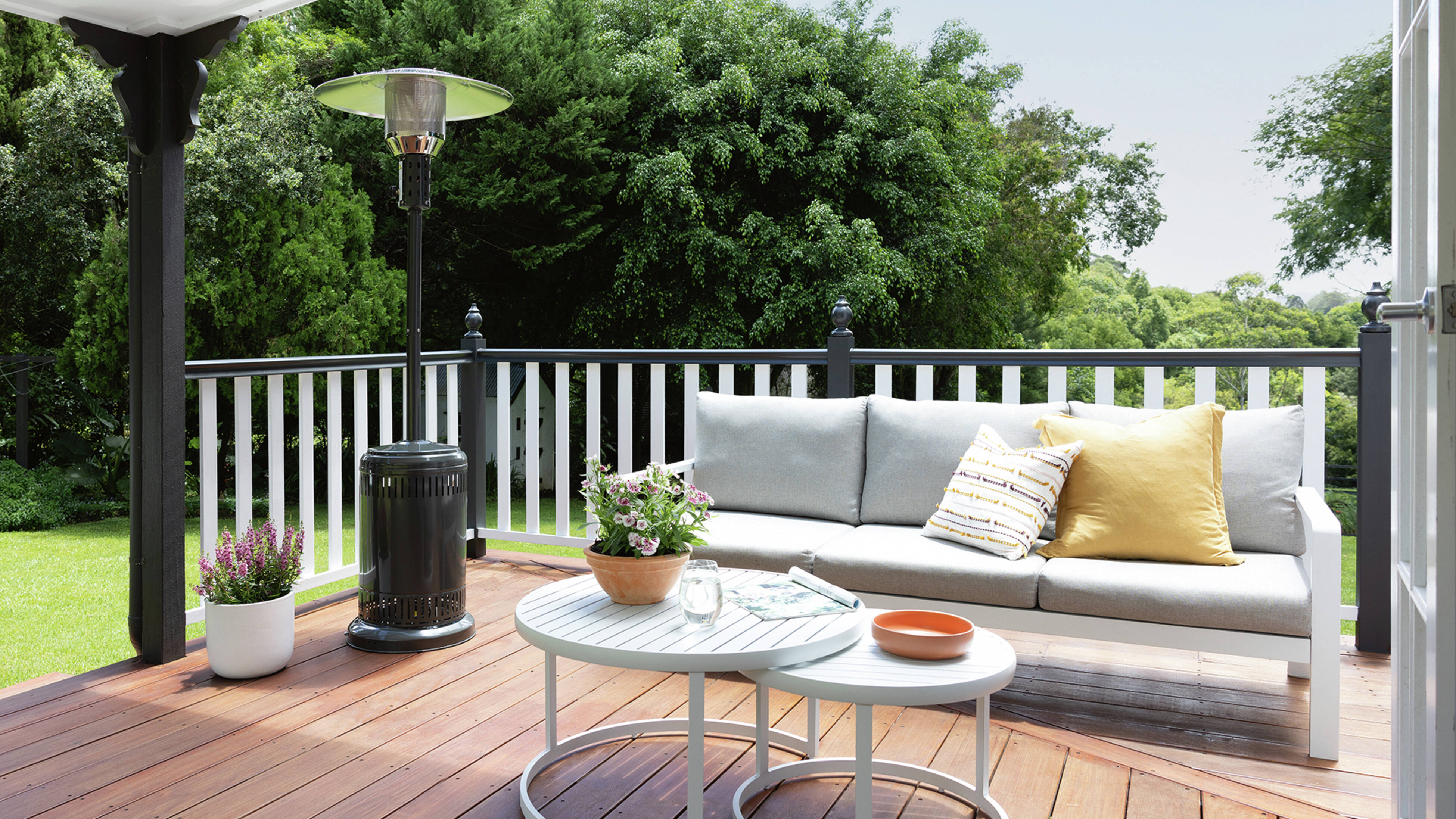Timber deck with a grey outdoor sofa, round white coffee table, and a tall gas patio outdoor heater beside a white railing with a lawn and trees in the background