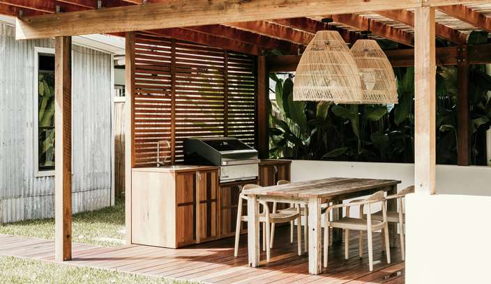 Outdoor timber pergola entertainment area featuring a covered dining space with a rustic wooden table and chairs, woven pendant lights, and a built-in barbecue with storage cabinetry. The deck overlooks a grassy lawn and is surrounded by tropical plants, creating a relaxed outdoor living and entertaining setting.