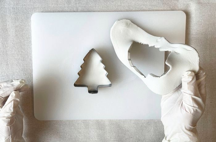 A white gloved hand holding white clay with a cut out shape of a Christmas tree, a cookie cutter with clay in a tree shape sits on a white chopping board.
