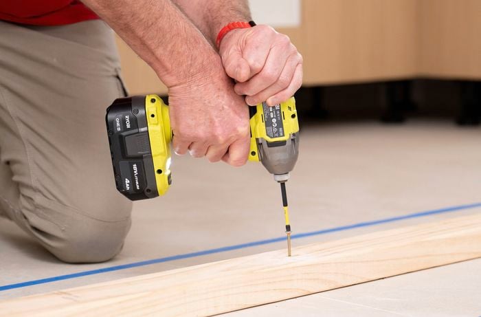 A person is using a power drill to fix the timber base to the floor.