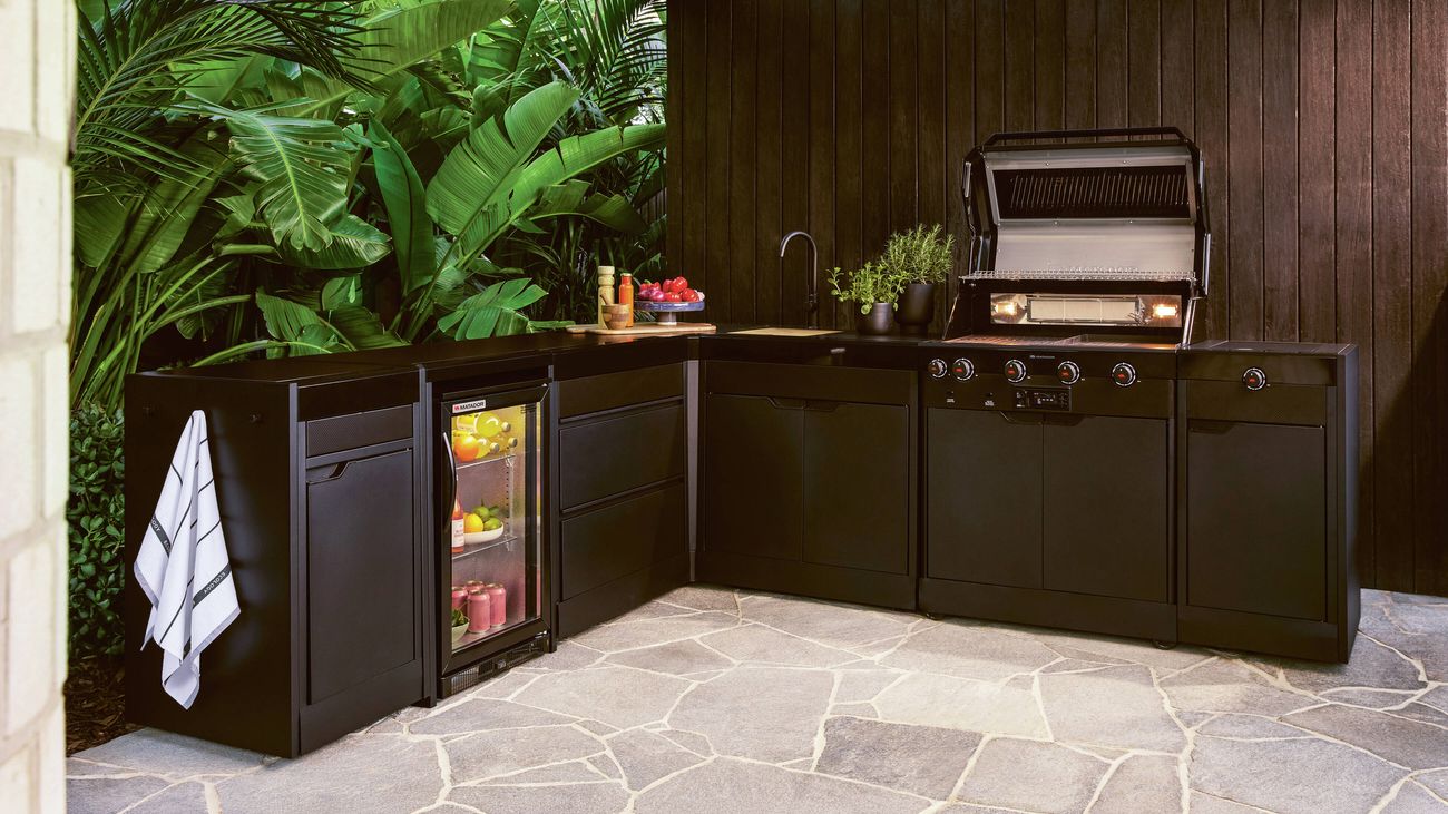 Outdoor kitchen with dark cabinetry, built-in BBQ, sink, and bar fridge, surrounded by lush greenery and wooden panelling.