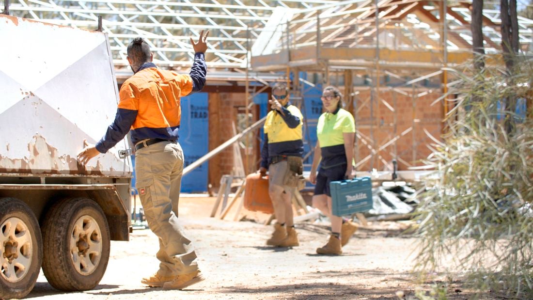 An image showing the back of a male tradie in orange hi-vis with one hand on the back of a work trailer and the other waving to two tradies – one male and one female – who are out of focus and carrying tools in boxes on a jobsite