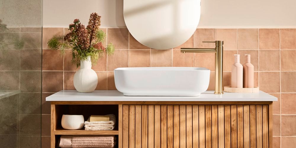 Timber vanity with open shelving, white basin and brushed gold tap set against warm terracotta tiles, styled with a vase of flowers and toiletries.