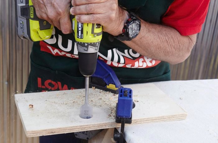 A bore spade drill bit drilling a wide hole through a plywood shelf to be threaded onto an ironing board storage rack