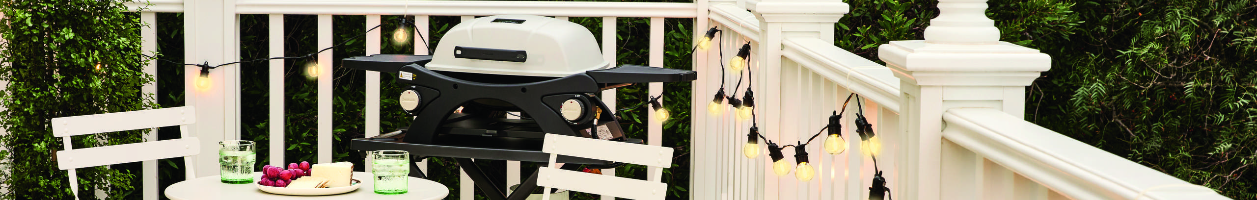 Outdoor entertaining area featuring a compact gas barbecue on a tiled deck, with a white bistro table set with drinks and snacks, white folding chairs, and warm string lights hanging along a white balustrade against a backdrop of dense greenery.