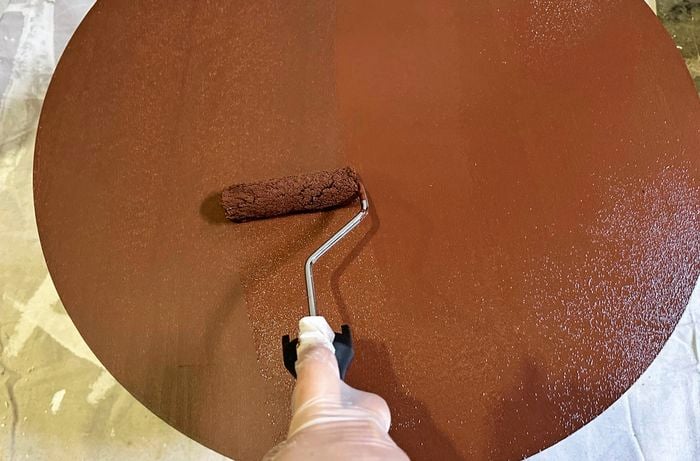 A gloved hand painting a round timber panel using a roller in brown