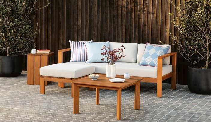 White and timber outdoor lounge setting on a paved area next to two pot plants with a fence behind it.