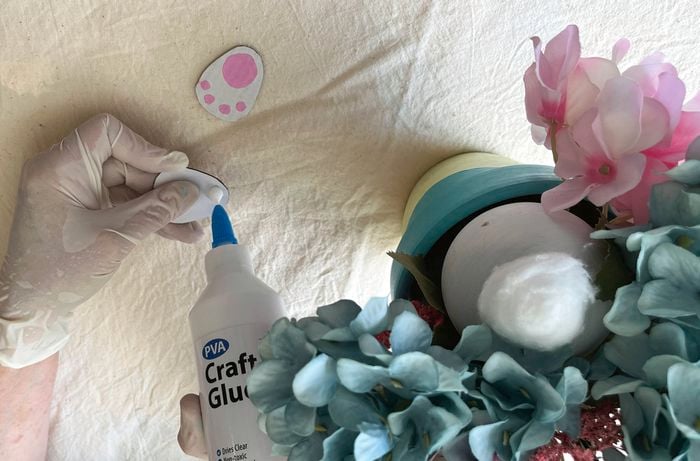 Hands wearing gloves insert artificial flowers into a block of floral foam, with pastel-coloured stems spread across a table.