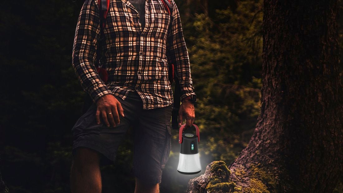 A person wearing a plaid shirt and backpack holding a lit camping lantern in a dark forest.