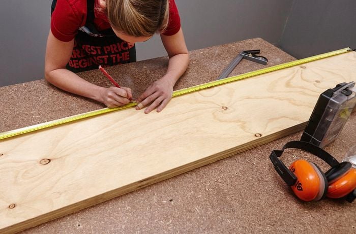 A person using a measuring tape to mark a measurement on a sheet of plywood