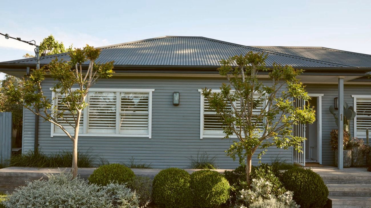 A grey house with a corrugated roof, white-shuttered windows, and a landscaped garden with trees and shrubs