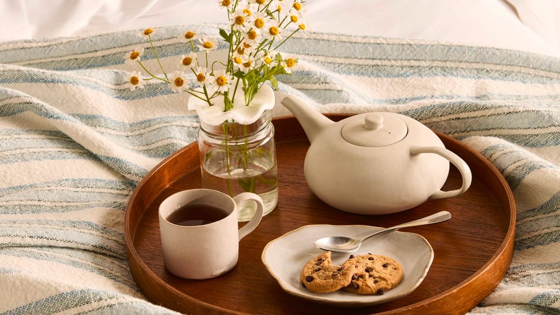 A wooden tray holding a glass vase with a flower frog holding chamomile flowers, a white mug of tea, a teapot and a plate with biscuits.