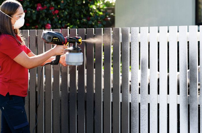 A person is applying paint evenly to a picket fence with a spray gun​​​ 