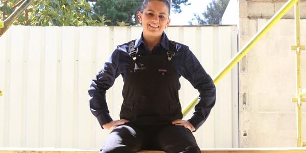 A female tradesperson wearing black Green Hip overalls sits on scaffolding on a job site.