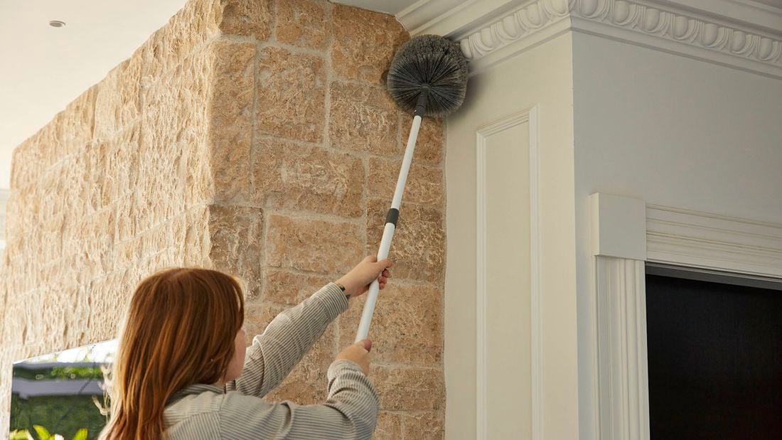  The back of a female using a brush with an extended handle to spring clean a ceiling corner 