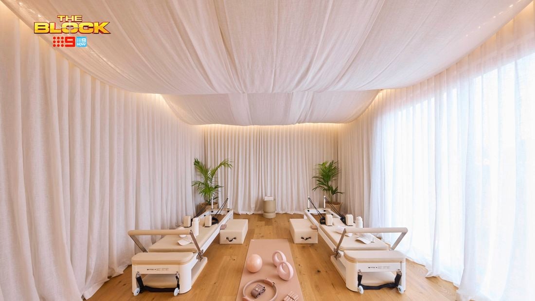 An elegantly designed Pilates studio with four reformer machines arranged symmetrically. The space features white draped curtains on the walls and ceiling, creating a serene ambiance. A pink exercise mat in the center holds pink fitness accessories including an exercise ball, resistance bands, and weights. Two potted plants sit at the far end, and the floor is light-colored wood. 