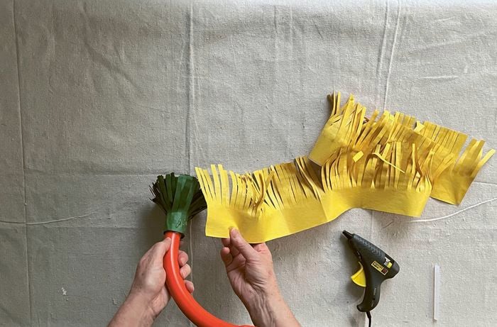 Hands attaching yellow fringed paper to an orange conduit bend with a green paper fringe, with a hot-glue gun nearby.