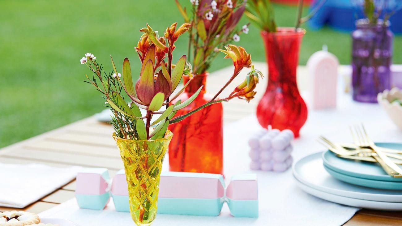 A close up image of colourful glass vases holding foliage and Christmas crackers and candles on the table.