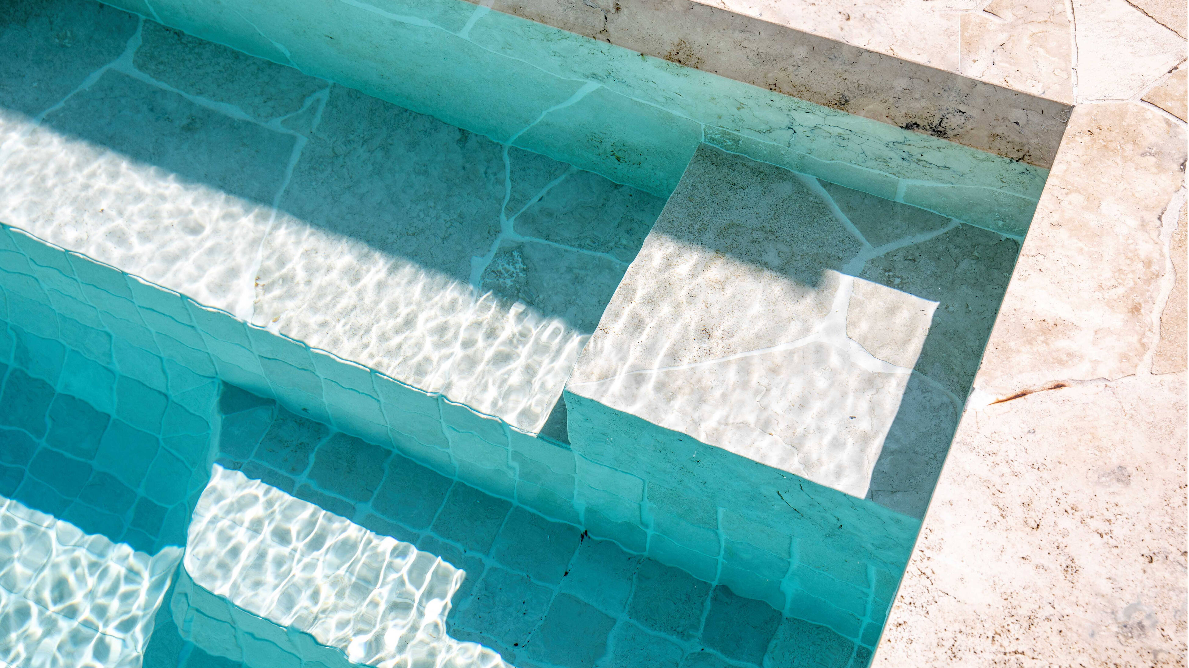 Close-up of a tiled swimming pool showing shallow steps under clear turquoise water and a light stone porcelain outdoor paving, with sunlight creating ripple patterns