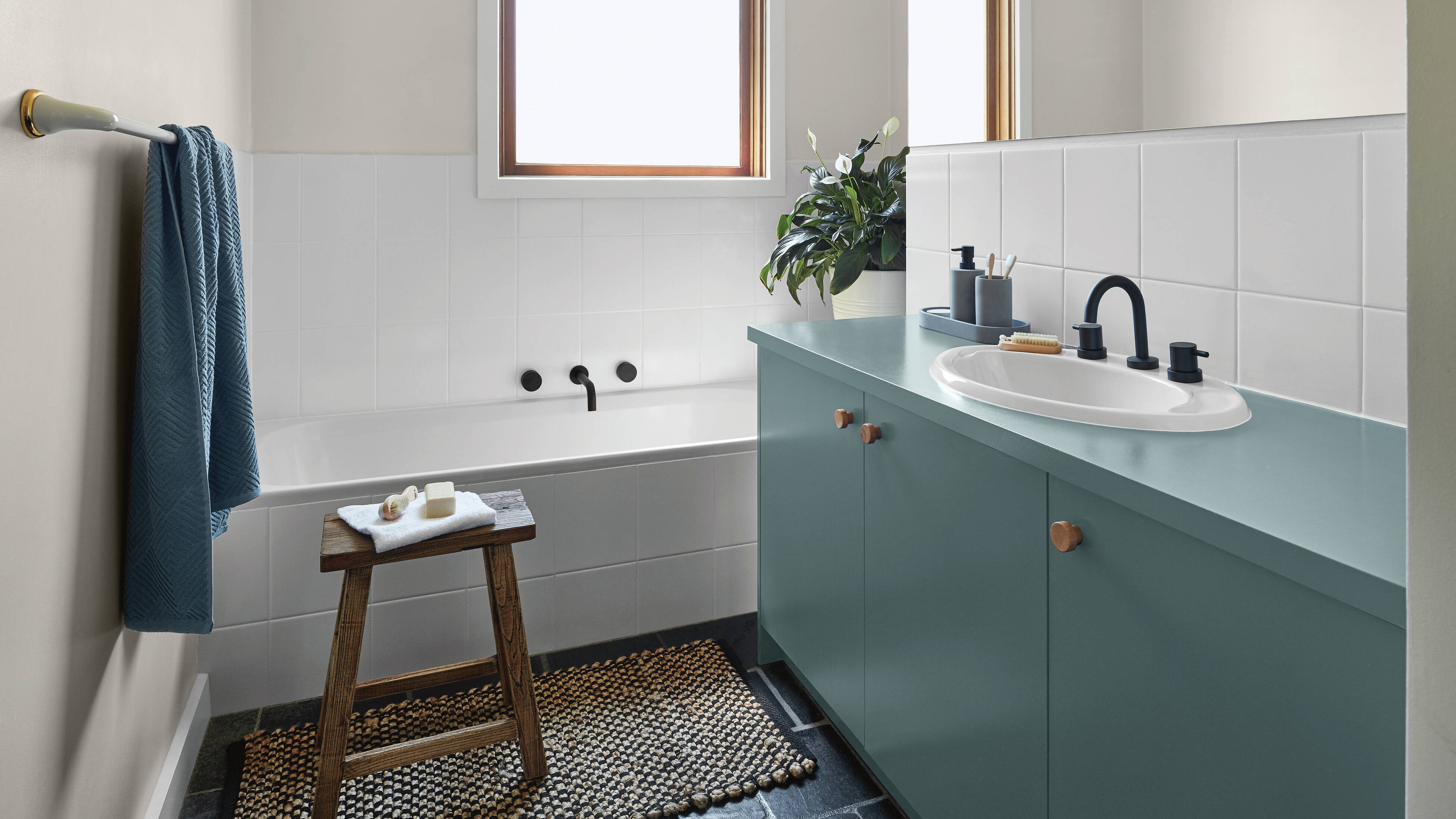 Bathroom with white tiled walls, a bathtub with black tapware, a teal vanity with a white basin and timber knobs, a wooden stool with soap, a textured rug on a dark floor, and a potted plant on the benchtop.