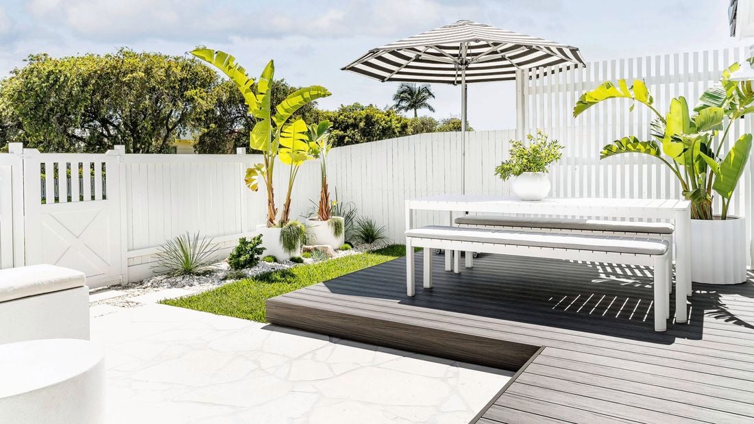A backyard design setting with a black and white striped sun umbrella, white outdoor setting and fence, plus greenery in white pots