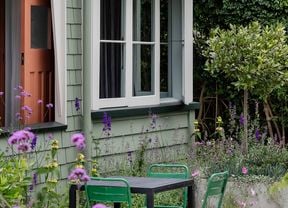 A weatherboard house painted with Dulux paint colours pale green with white trims. Small courtyard with a black table and four green chairs and purple flowers.