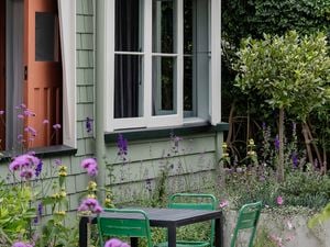 A weatherboard house painted with Dulux paint colours pale green with white trims. Small courtyard with a black table and four green chairs and purple flowers.