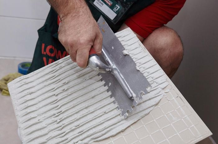 Person running notched trowel through glue on the back of a new tile.