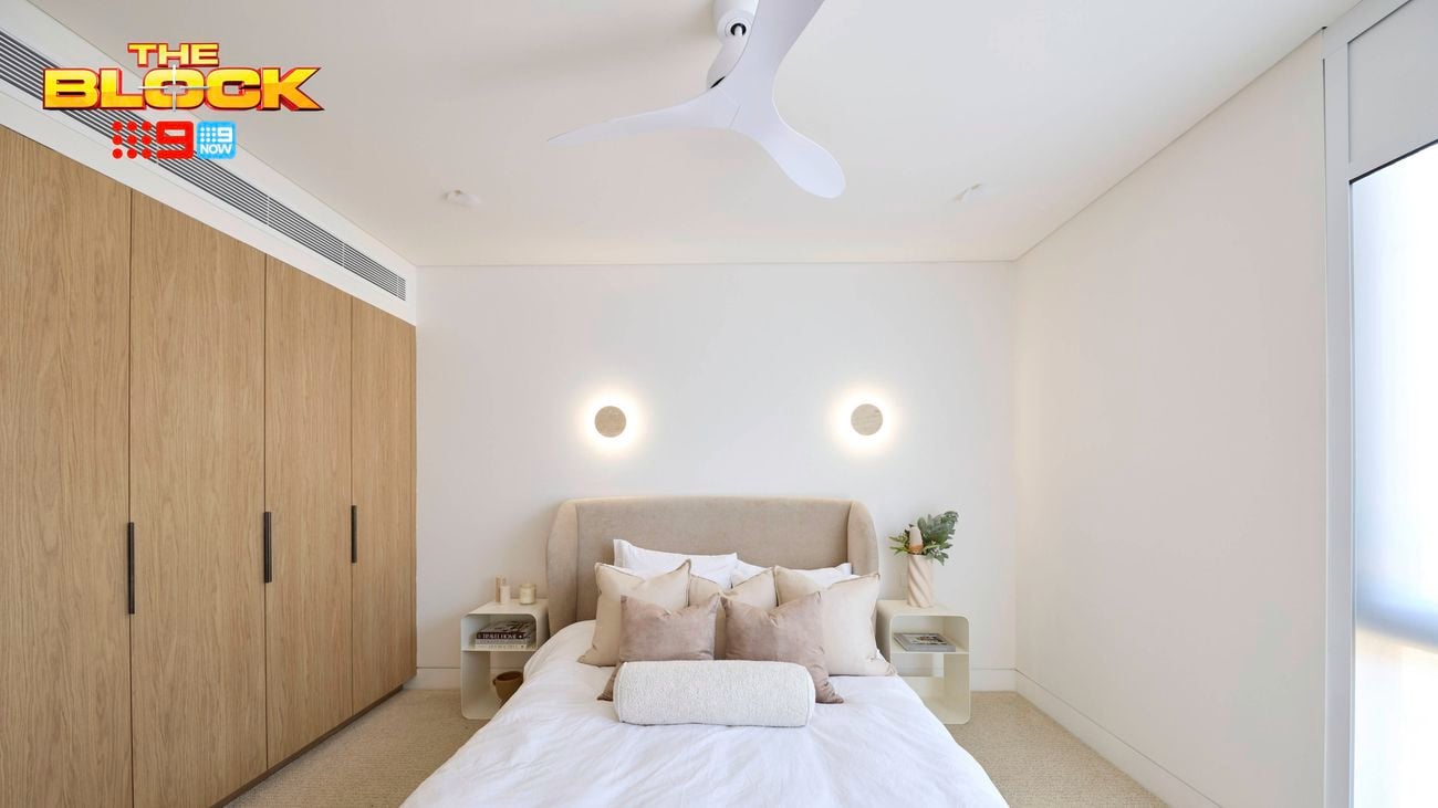 A furnished bedroom with a bed in it, two side tables, a wardrobe and carpet on the floor and The Block logo on the image.