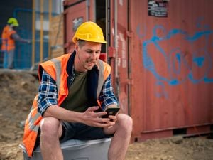 Construction worker in high-visibility vest and yellow hard hat sitting beside a shipping container, using a smartphone, with another worker and scaffolding in the background.
