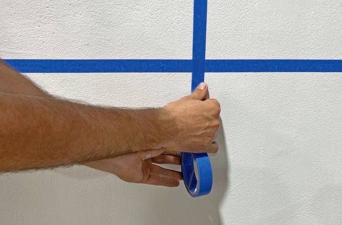 Blue painter’s tape making a cross on a white wall to start colour block design, with the roll of tape held by two hands.