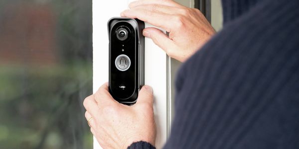 Close-up of a person installing a smart video doorbell on a white door frame, with the camera lens and doorbell button visible beside a glass panel.
