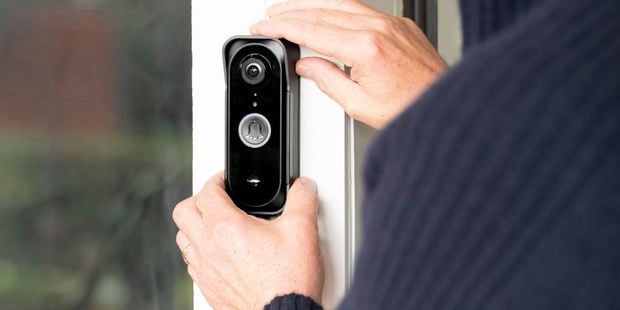 Close-up of a person installing a smart video doorbell on a white door frame, with the camera lens and doorbell button visible beside a glass panel.