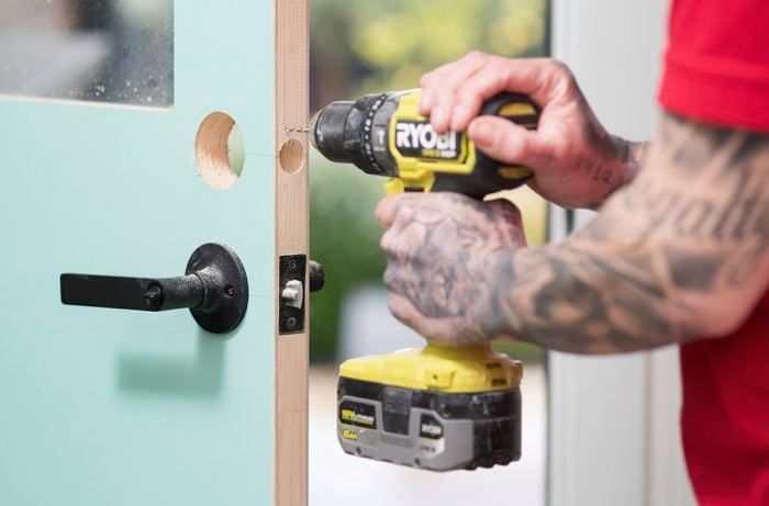 A person is using a drill to create a pilot hole on the edge of the door for a latch installation