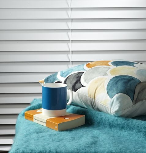 Close up of mug, book and cushion in front of a window with white venetian blinds