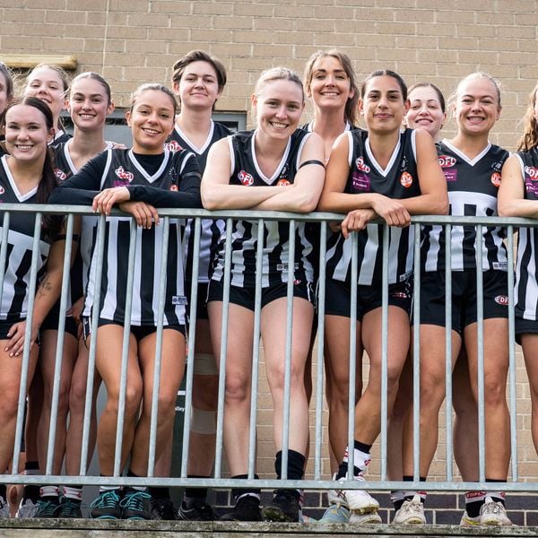 A women’s sports team in matching black uniforms posing together behind a metal railing outdoors, smiling at the camera.