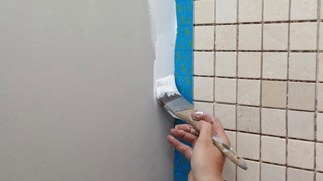 A person painting down the edge of a plasterboard wall next to a tiled wall with masking tape