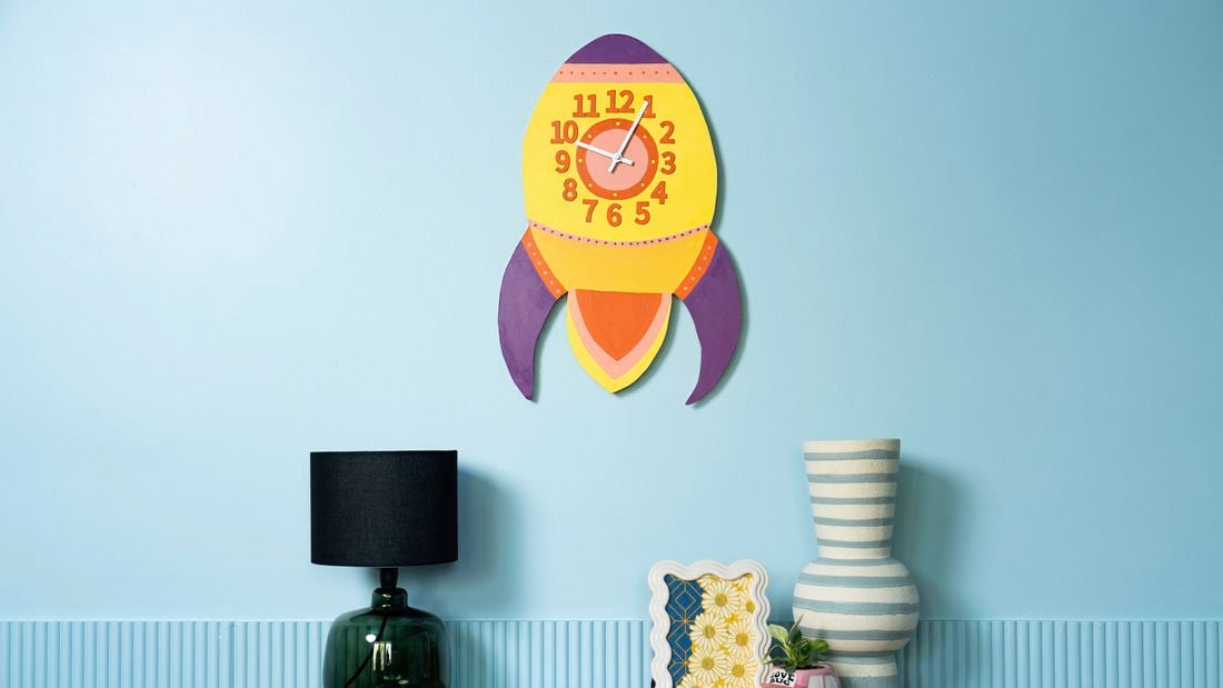 A colourful retro rocket wall clock displayed on a blue wall above a wooden console table with modern decorative pieces.