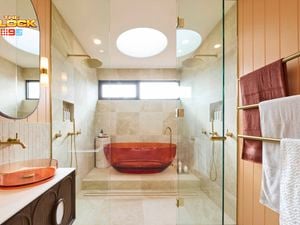 Modern bathroom with amber tub in glass shower, beige tiles, dual overhead showerheads, amber basin with round mirror, and gold towel rack. 