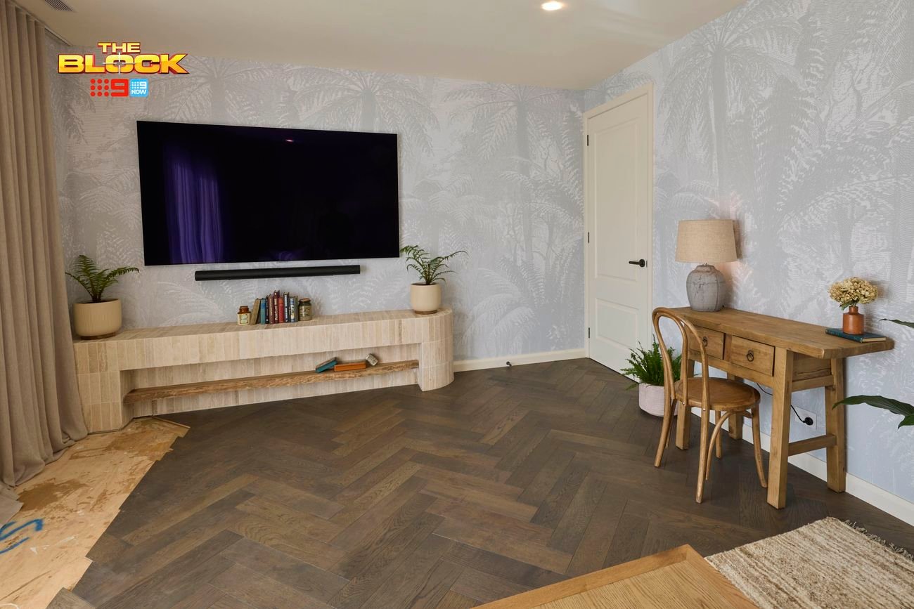 A modern living room featuring a wall-mounted flat-screen TV above a wooden shelf with books and decorative items. To the right, a wooden desk with a chair, lamp, and plants sits against light-patterned wallpaper. The floor has a herringbone design.