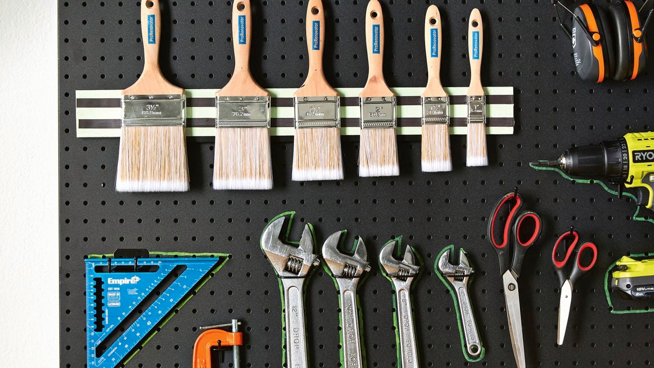  Pegboard above a wooden benchtop with neatly arranged tools: paintbrushes, screwdrivers, spanners, scissors, a cordless drill, rafter square, a G‑clamp, and a spirit level.