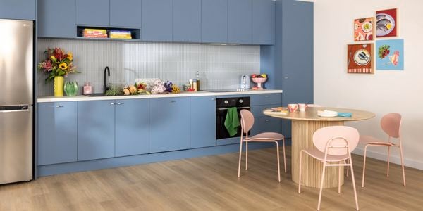 Modern kitchen with blue cabinets, light wooden floor, stainless steel fridge, countertop with flowers, fruits, and appliances, round wooden dining table with four pink chairs, and four colorful food-themed framed pictures on the wall.