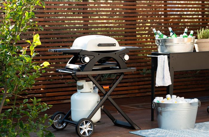 Portable gas barbecue on an outdoor deck beside a drinks cart and cooler tubs.