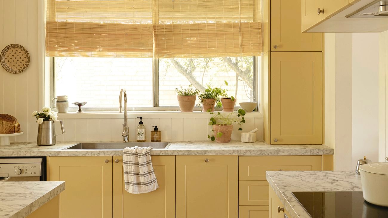 Yellow kitchen with panelled cabinets, marble‑look benchtops and light timber flooring. A wide window with bamboo blinds above the sink holds terracotta herb pots.