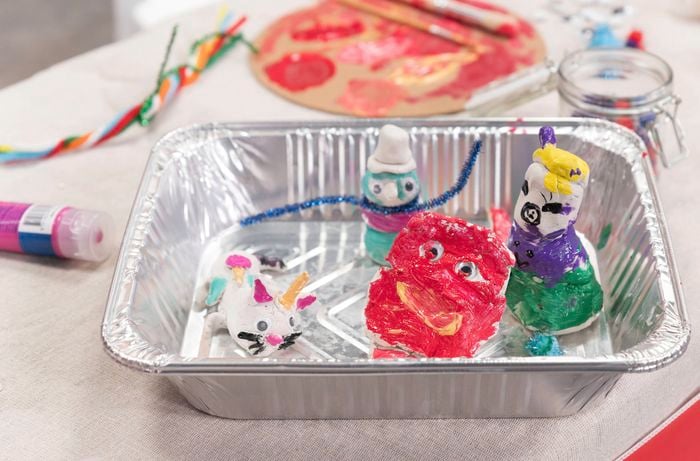 Painted clay figures with googly eyes and vibrant designs are displayed in a BBQ tray on a craft table