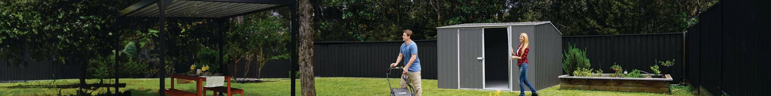 Man with a lawn mower and a woman closing a door to a shed in a backyard.