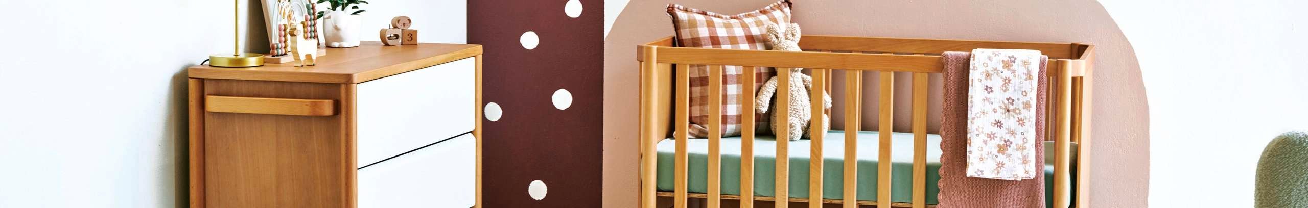 White walled bedroom with a wooden cot, dresser and green rocking chair. The walls are painted with patterns of a dusty colour palette and toys are strewn on the floor.